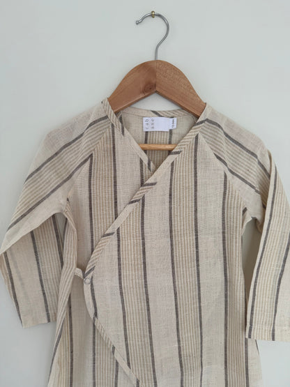 The APRÈS WRAP natural stripe by cabane childrenswear is a beige long-sleeve baby kimono with thin gray and tan stripes, made from breathable fabric, displayed on a wooden hanger against a light-colored wall.