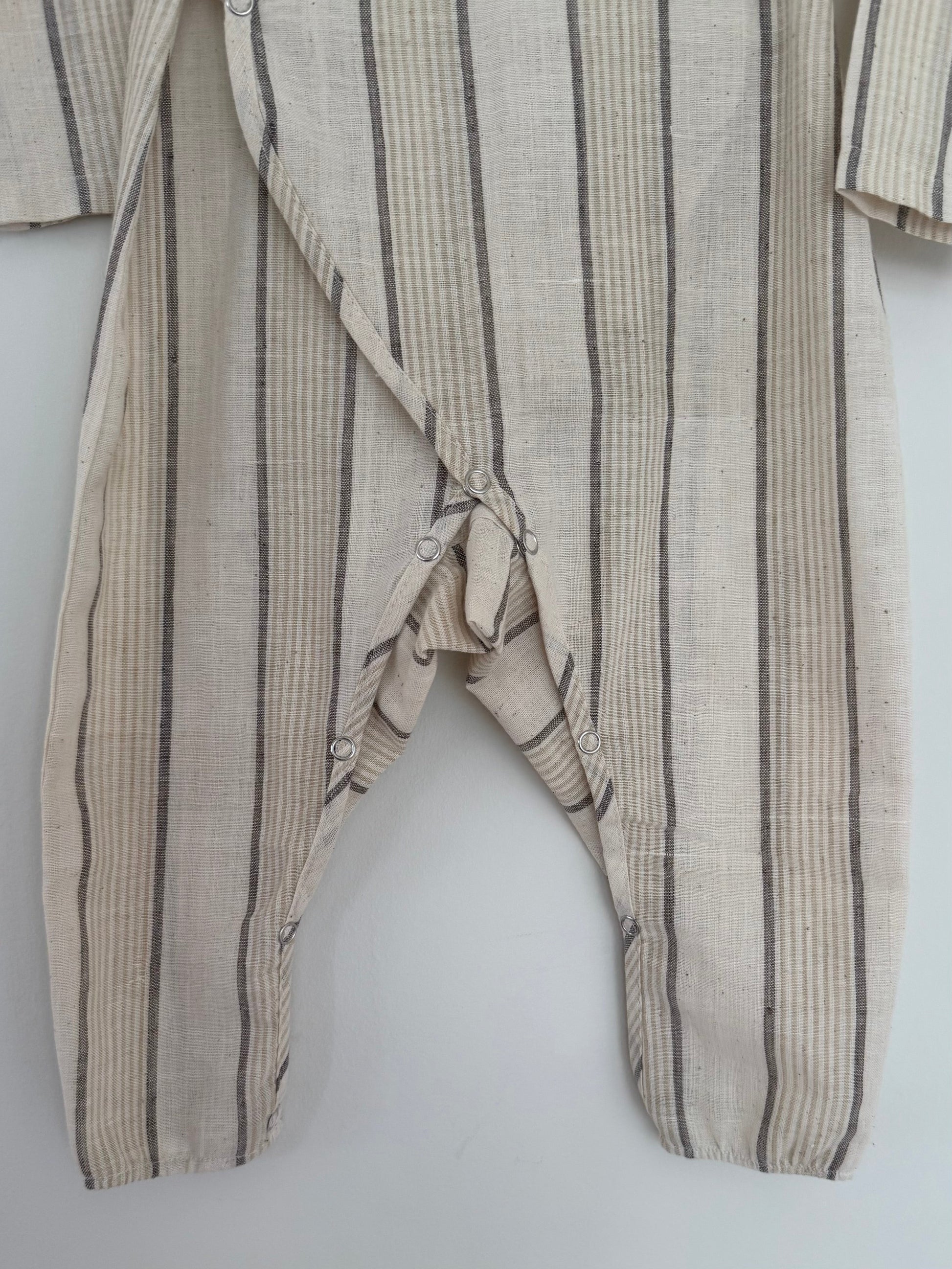 The cabane childrenswear APRÈS WRAP natural stripe romper in beige features black and gray vertical stripes, breathable fabric, and snap buttons along the inseam and one leg; shown hanging against a light background.