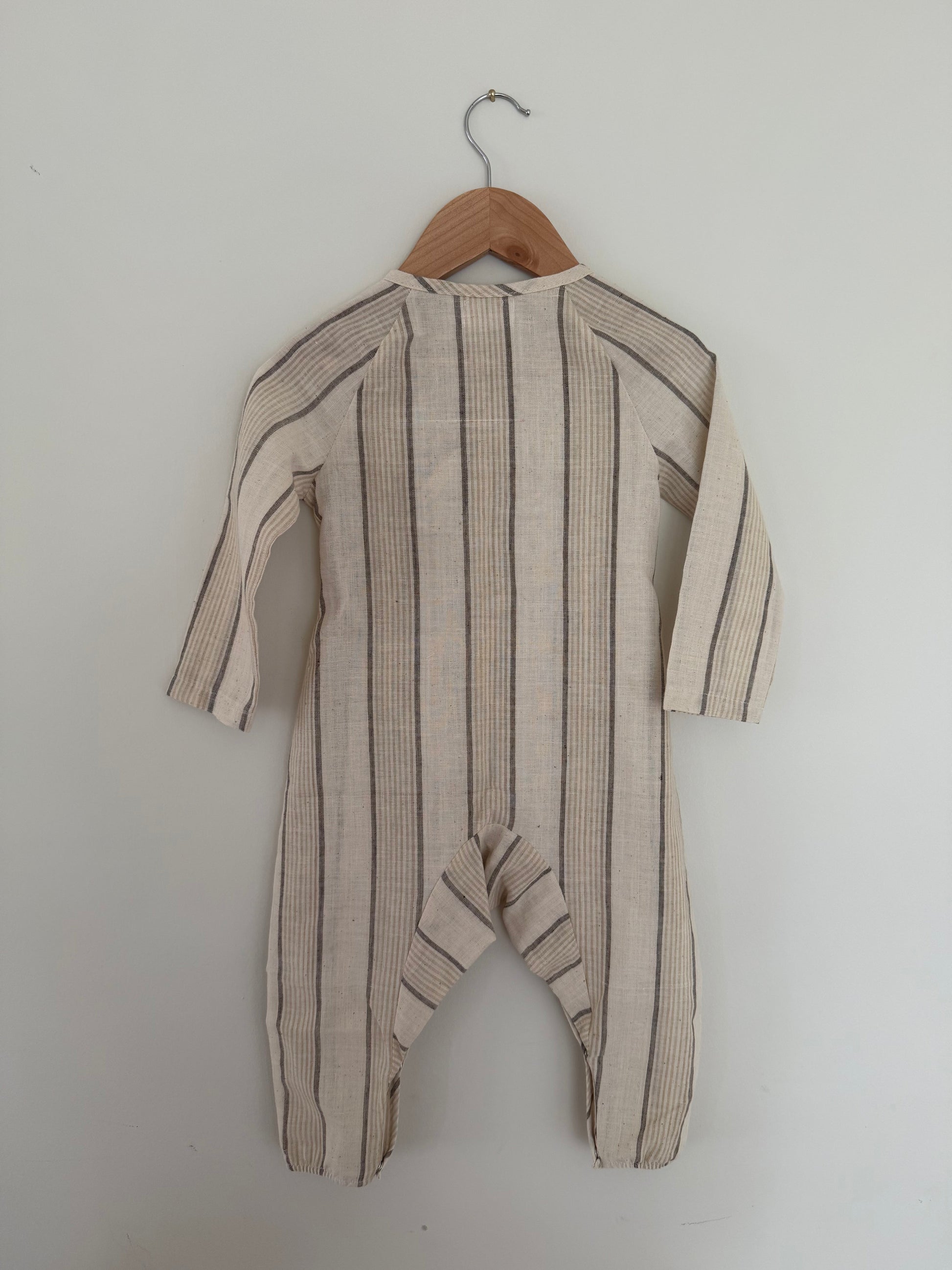 The APRÈS WRAP natural stripe by cabane childrenswear—a beige, long-sleeved baby romper with vertical black stripes—hangs on a wooden hanger against a plain white wall.