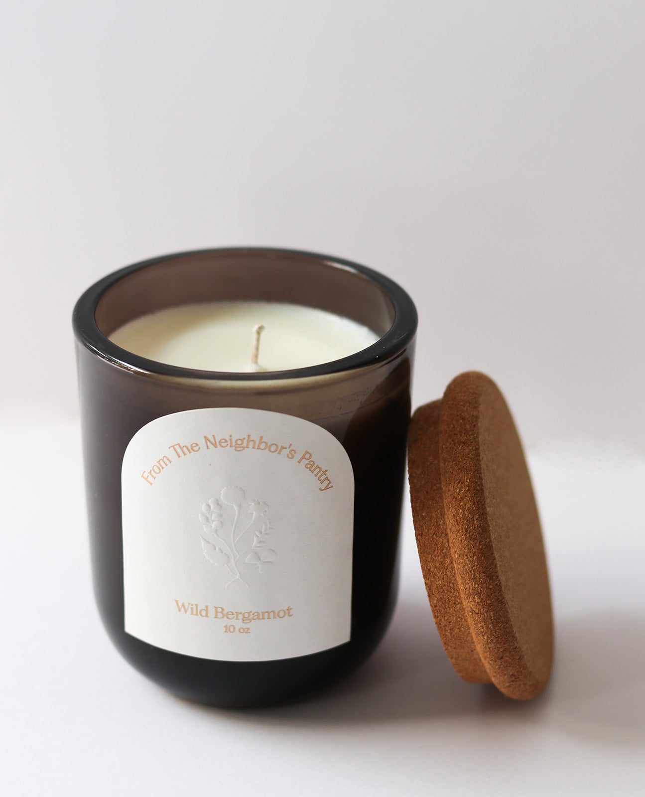 A brown glass Wild Bergamot Tallow Candle by My Neighbor’s Tallow features beeswax, an embossed plant design, and “From The Neighbor's Pantry” text. It has a single wick and comes with a cork lid, shown on a white background.