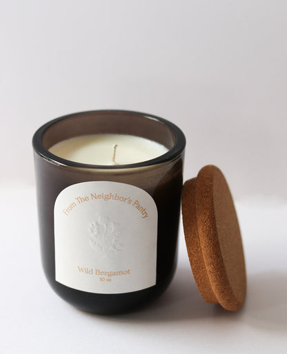 A brown glass Wild Bergamot Tallow Candle by My Neighbor’s Tallow features beeswax, an embossed plant design, and “From The Neighbor's Pantry” text. It has a single wick and comes with a cork lid, shown on a white background.