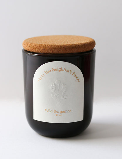 A dark glass jar candle with a cork lid and a white label featuring "My Neighbor’s Tallow" and floral art, scented “Wild Bergamot Tallow Candle,” 10 oz, sits against a plain background.