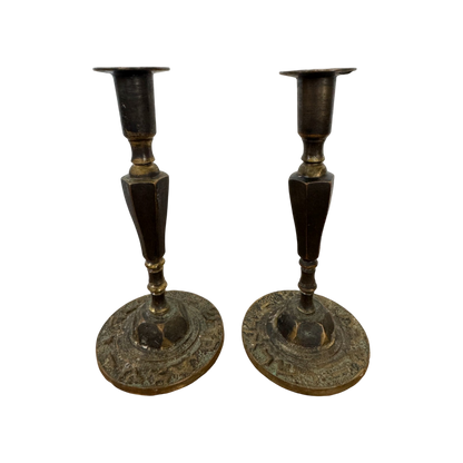 A pair of Shoppe Maison Vintage Shabbat Candle Holders with tall, tapered shafts and wide, circular bases featuring intricate etched designs, set against a black background. These brass candlesticks show signs of tarnish, adding to their antique appearance.