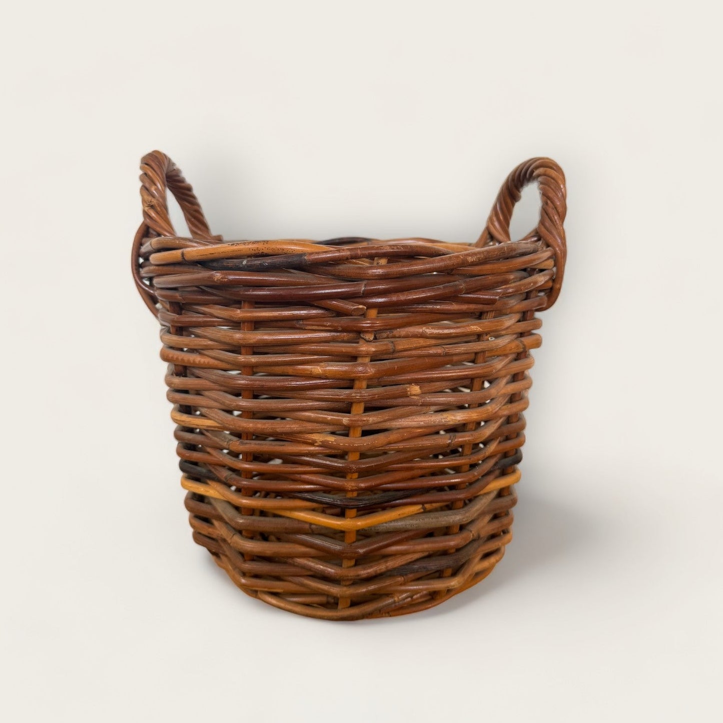 The Vintage Woven Basket with Handles from Shoppe Maison is a rustic, round wicker basket featuring two handles on either side. Made of interwoven natural wood fibers, this vintage basket showcases a blend of various shades of brown. The plain, light-colored background accentuates the basket's natural texture and pattern.