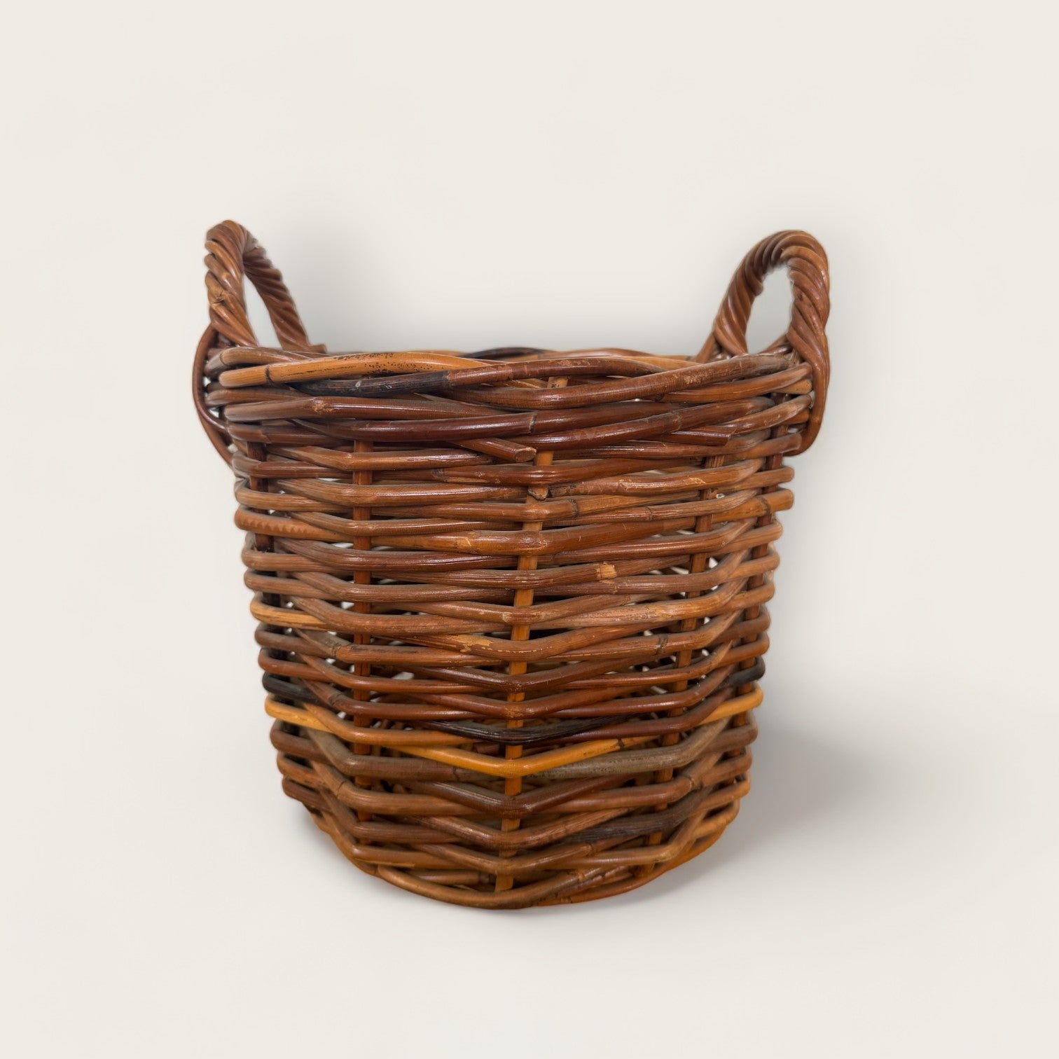 The Vintage Woven Basket with Handles from Shoppe Maison is a rustic, round wicker basket featuring two handles on either side. Made of interwoven natural wood fibers, this vintage basket showcases a blend of various shades of brown. The plain, light-colored background accentuates the basket's natural texture and pattern.