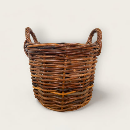 The Vintage Woven Basket with Handles from Shoppe Maison is a rustic, round wicker basket featuring two handles on either side. Made of interwoven natural wood fibers, this vintage basket showcases a blend of various shades of brown. The plain, light-colored background accentuates the basket's natural texture and pattern.