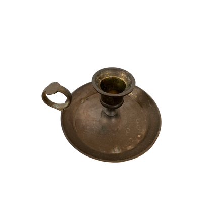 The Brass Candle Holder II by Shoppe Maison features a vintage design with a circular base and a finger loop handle on one side. The tarnished metal reflects its age and wear, adding to its antique charm. A drip tray encircles the empty candle socket, and the black background accentuates the timeless elegance of this piece.