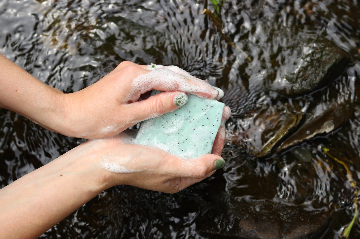 Hands with green-painted nails lather a light green Blue Tansy & Raw Honey Tallow Soap bar by My Neighbor’s Tallow in clear water over outdoor rocks.