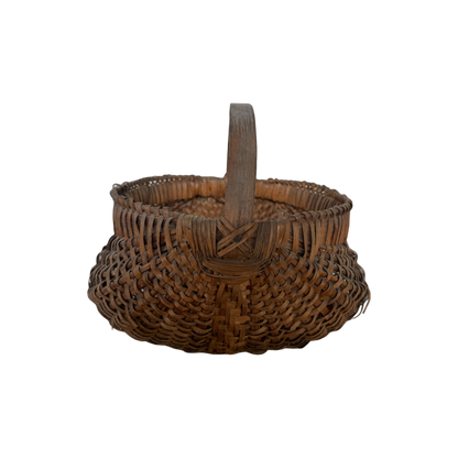 The Antique Gather Basket II by Shoppe Maison features a sturdy handle, rounded bottom, and decorative pattern. Reminiscent of antique buttocks baskets, this woven wicker basket appears empty and is displayed against a black background. The detailed and intricate weaving technique gives it a rustic, handcrafted feel perfect for decorative purposes.