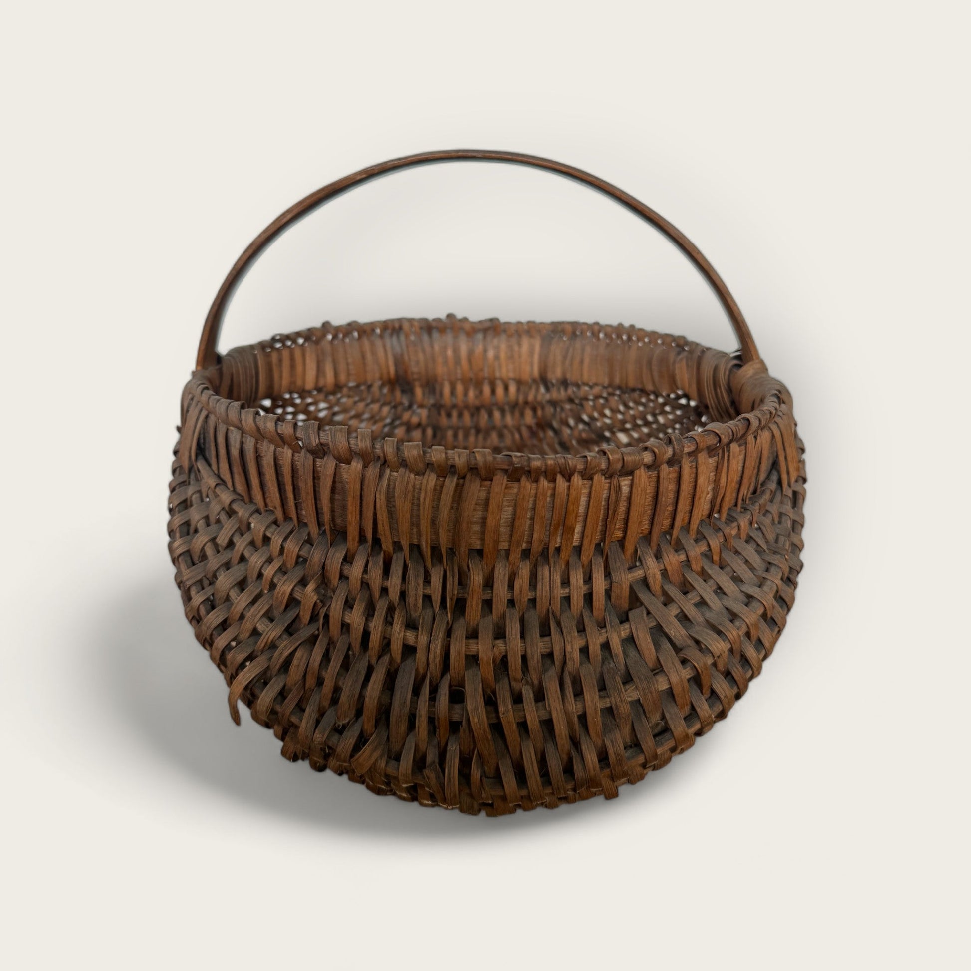 The Antique Gather Basket II by Shoppe Maison features a round, brown wicker design with a single curved handle extending from one side to the other. The basket's tight and consistent weave displays a traditional woven pattern, making it perfect for decorative purposes. The plain background allows this vintage piece to be clearly seen.