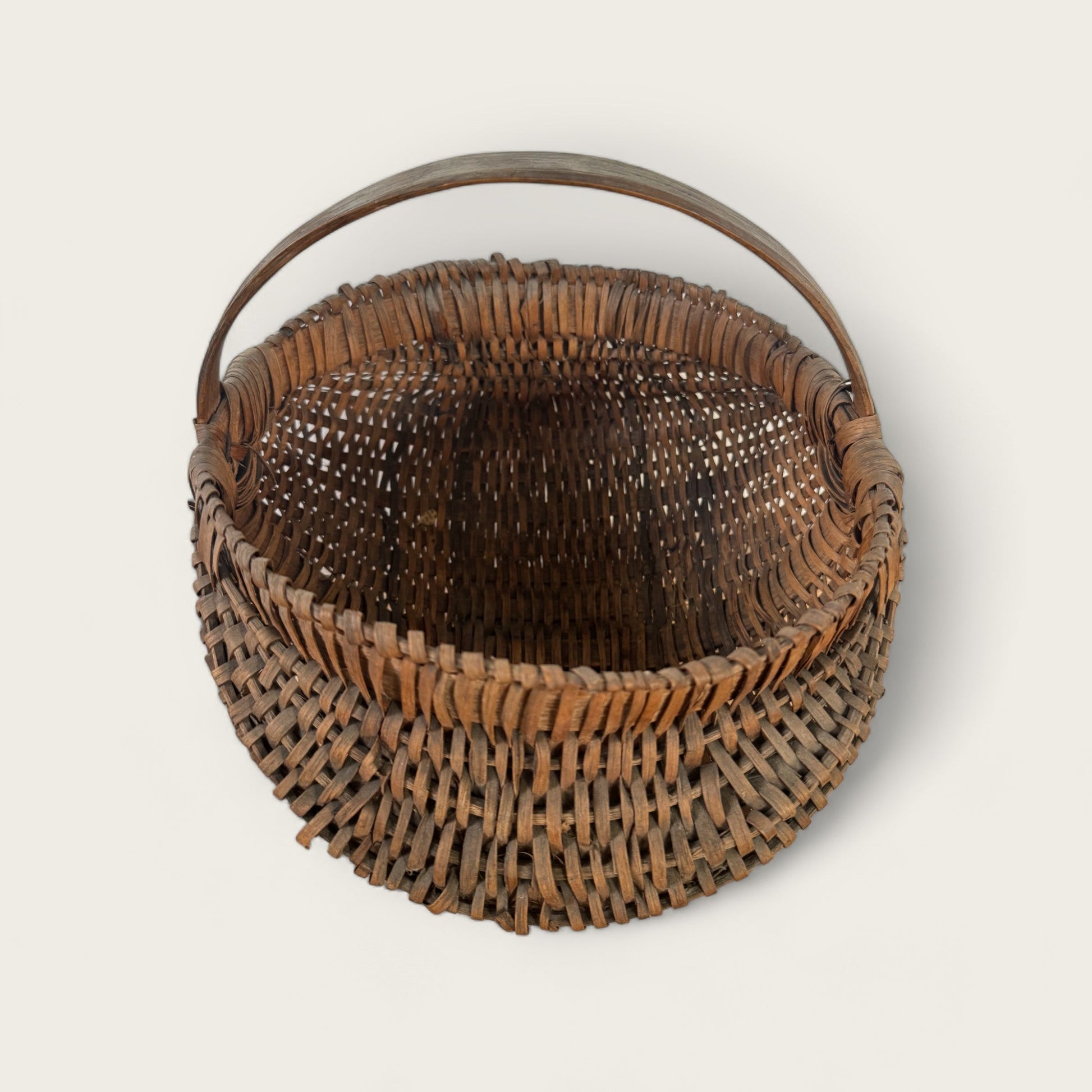 An empty, round, open-top *Antique Gather Basket II* from *Shoppe Maison*, complete with a sturdy handle, is showcased. Made from interlaced wooden or bamboo strips, it boasts a rustic, natural brown finish. This vintage item is practical for carrying or storing various articles and doubles as an excellent decorative piece.