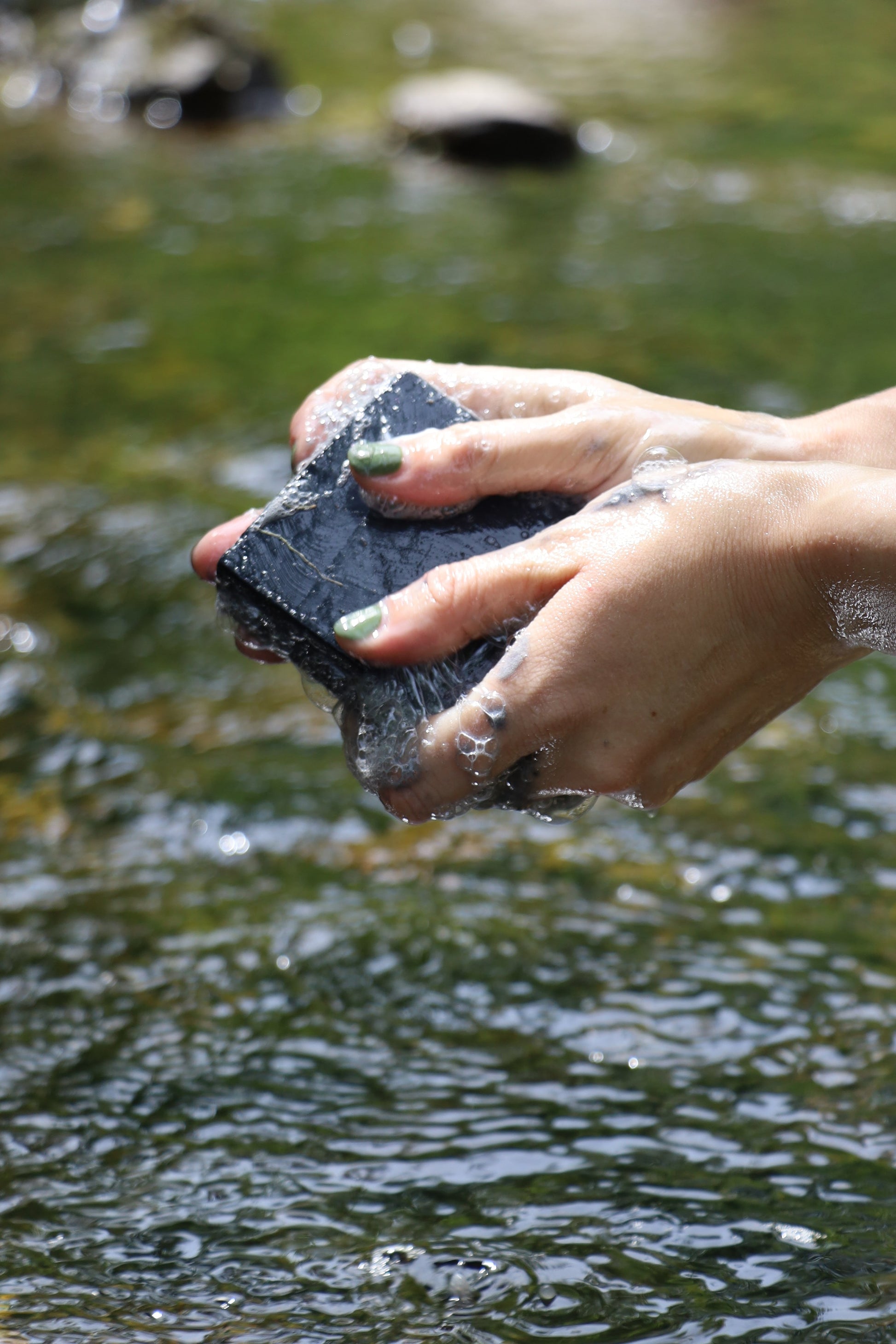 Hands with green-painted nails wash a dark gray stone in clear, bubbling river water using My Neighbor’s Tallow Charcoal Tallow Soap, set against a blurred green background.