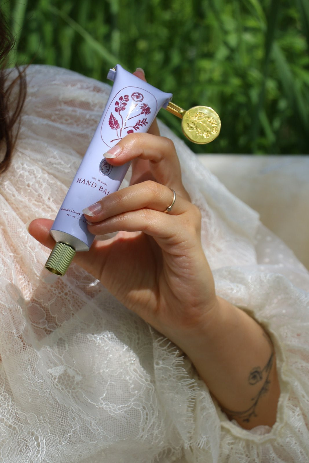 A person in a white lace garment holds a My Neighbor’s Tallow Manuka Honey & Lavender hand balm tube, featuring floral illustrations and a gold seal, with soft greenery blurred in the background.