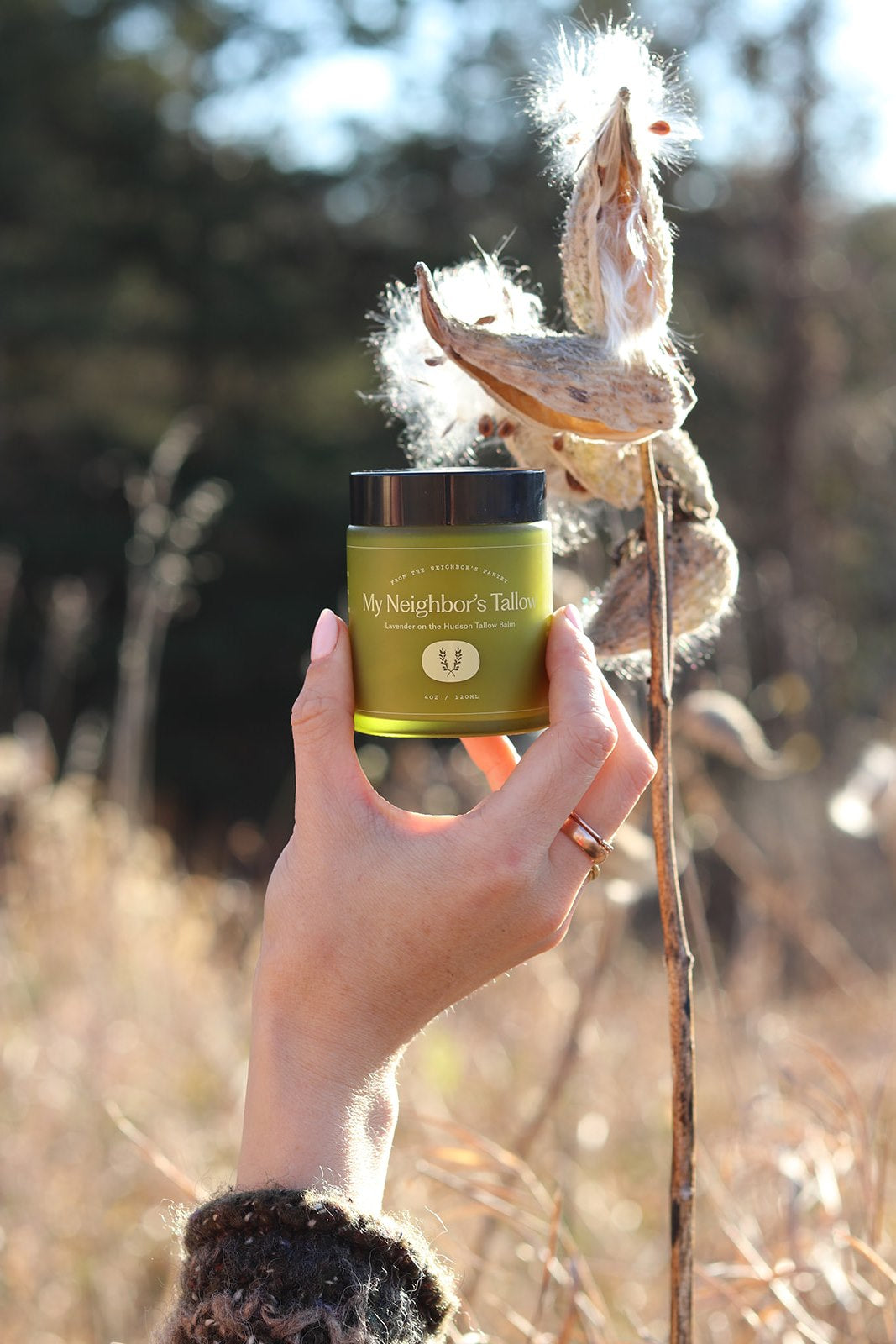 A hand with a ring holds up a green jar of “Lavender on the Hudson - Tallow Balm” by My Neighbor’s Tallow outdoors, with dry plants and soft sunlight in the background.
