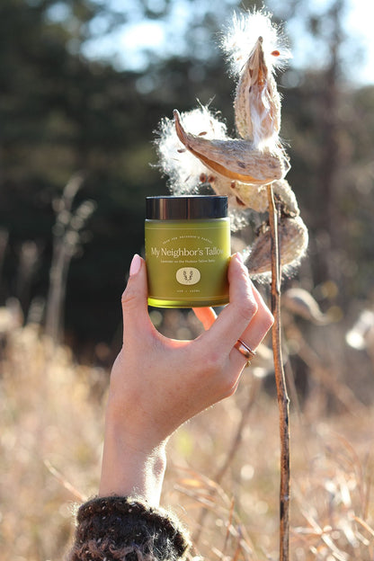 A hand with a ring holds up a green jar of “Lavender on the Hudson - Tallow Balm” by My Neighbor’s Tallow outdoors, with dry plants and soft sunlight in the background.