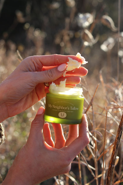 A person scoops Lavender on the Hudson - Tallow Balm by My Neighbor’s Tallow from a green jar while standing outdoors among dry grass and sunlight.