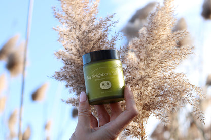 A hand holds up a green jar labeled "My Neighbor’s Tallow Wild Bergamot - Tallow Balm" with tall beige pampas grass and a clear blue sky in the background.