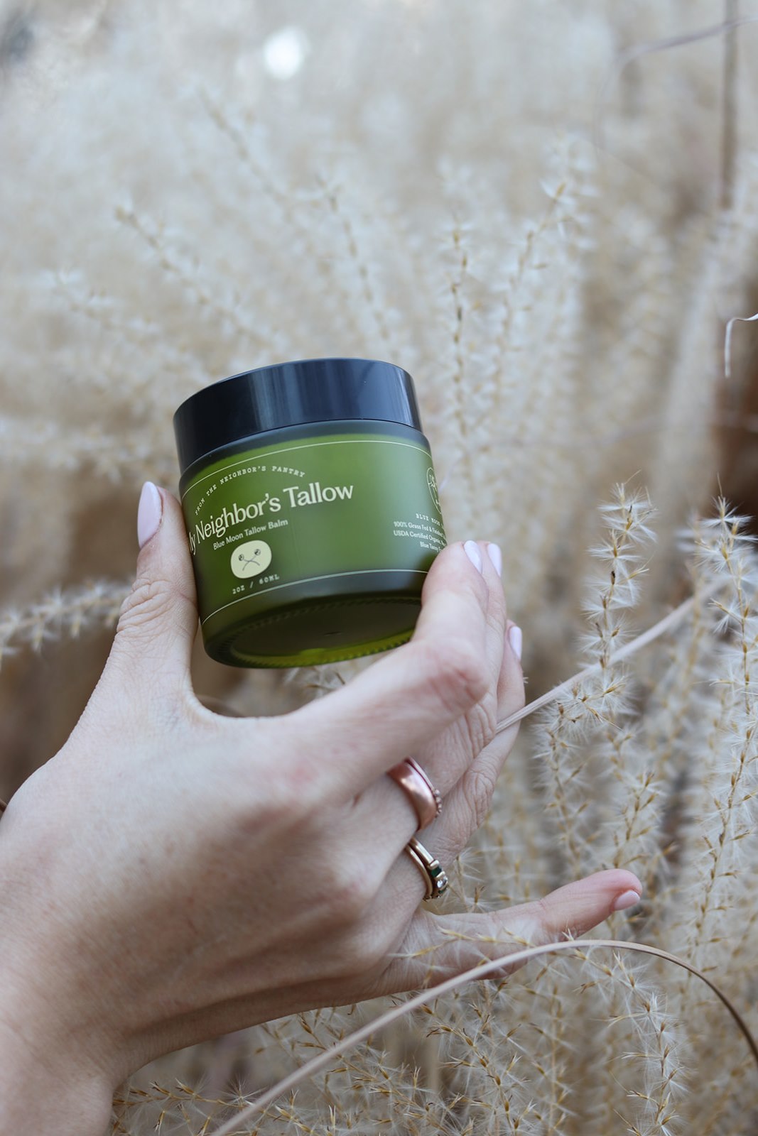 A hand with rings holds a green jar labeled "Blue Moon Face Balm" by My Neighbor’s Tallow—a moisturizer renowned for enhancing skin elasticity—set against a background of dried grass or wheat.