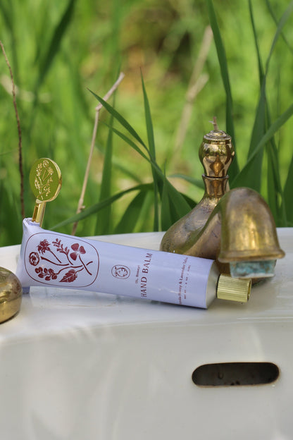 A tube of My Neighbor’s Tallow Manuka Honey & Lavender hand balm with a gold cap and floral design sits on white ceramic outdoors, surrounded by brass accents and lush green grass in the background.