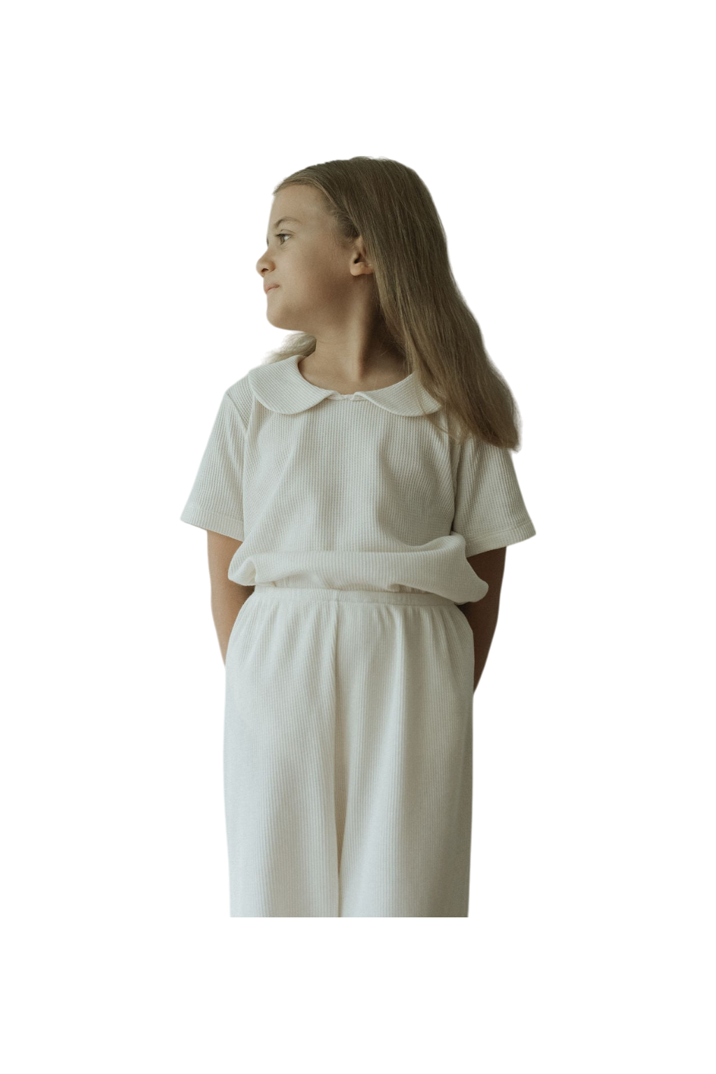 A young girl with long hair stands looking to the side in a SCHOOLHOUSE TEE waffle by cabane childrenswear, embodying classic looks with a collared top and relaxed pants. The plain, neutral background highlights her serene expression, encapsulating the essence of the brand.