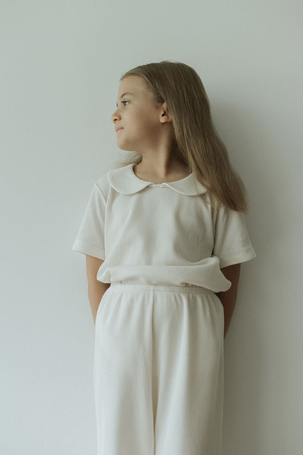 A young girl stands against a white wall, gazing left and embodying receptivity. She wears cabane childrenswear's SCHOOLHOUSE TEE waffle, an off-white short-sleeved top with long pants. Her hair flows freely as her hands are tucked behind her back in a minimalist, serene setting.