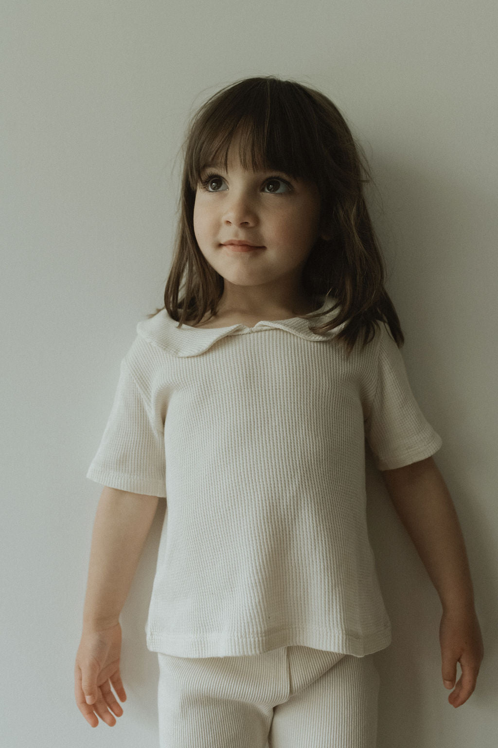 A brown-haired child stands against a white background, embodying classic charm in a ribbed beige outfit. The attire features the SCHOOLHOUSE TEE waffle and matching pants by cabane childrenswear. They gaze slightly upward with a soft smile, exuding warmth and receptivity.