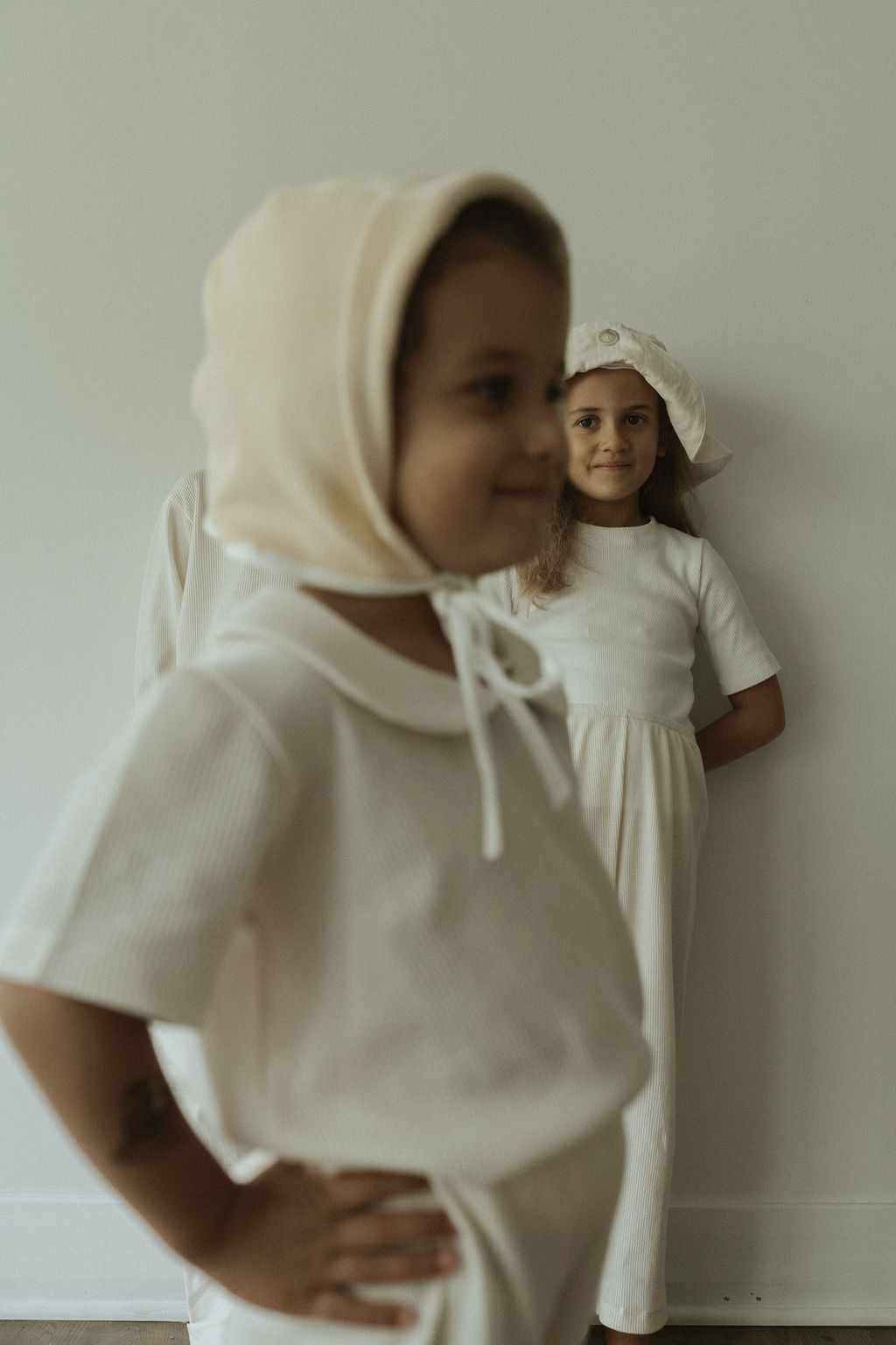 Two children embody classic style in white outfits and hats. The front child beams with a bonnet, while the one behind wears a cap and stands with hands on hips against a plain wall, echoing cabane childrenswear's timeless charm with the SCHOOLHOUSE TEE waffle.