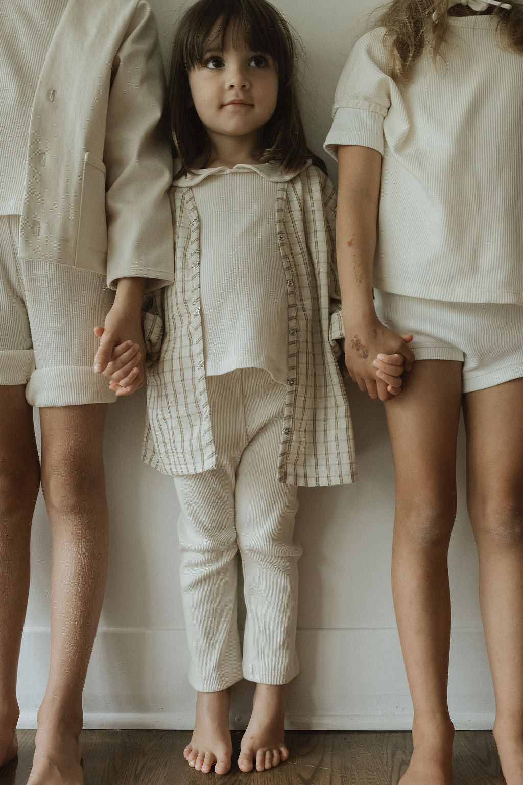 Three young children stand barefoot against a wall, holding hands. The two side children wear cabane childrenswear FAVORIS rib pants, with only their lower halves visible. The center child wears a plaid shirt and cream outfit.