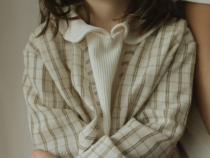 A child wears the cabane childrenswear TAKE CARE TOP in olive check, standing with arms crossed and partially leaning against someone out of frame. The child's face is partially visible.