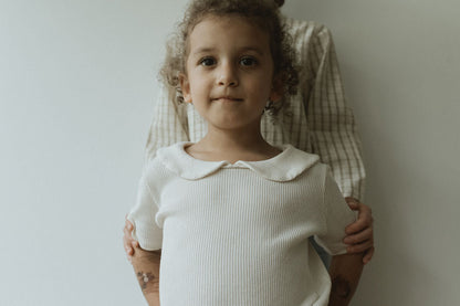 A young child with curly hair poses in a SCHOOLHOUSE TEE waffle by cabane childrenswear, embodying classic looks. They stand before an adult in a plaid shirt with hands resting gently on the child's shoulders, all set against a plain wall that highlights their connection and timeless appeal.