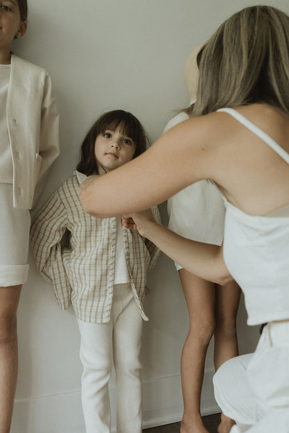 A woman helps a young girl button a plaid cotton shirt while two other children, dressed in cabane childrenswear FAVORIS rib pants, stand nearby in neutral tones against a plain wall.