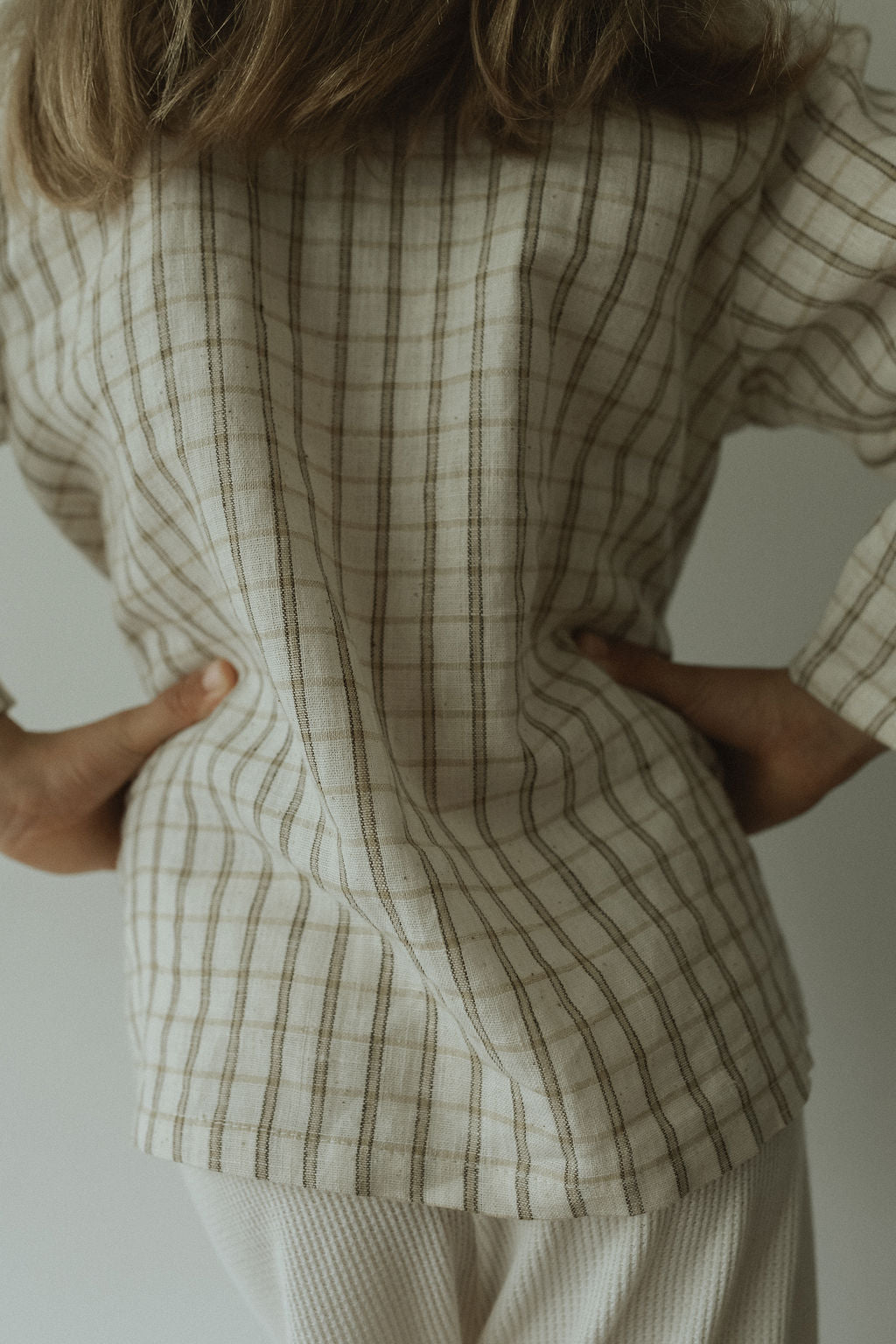 A person stands with hands on hips, seen from behind, wearing the cabane childrenswear TAKE CARE TOP in olive check—an ethically handmade long-sleeve shirt—paired with light-colored pants.