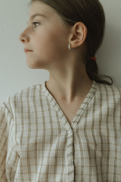 A young girl with light skin and brown hair in a ponytail looks to the side, wearing the cabane childrenswear TAKE CARE TOP in olive check and a small hoop earring. The background is plain and light-colored.