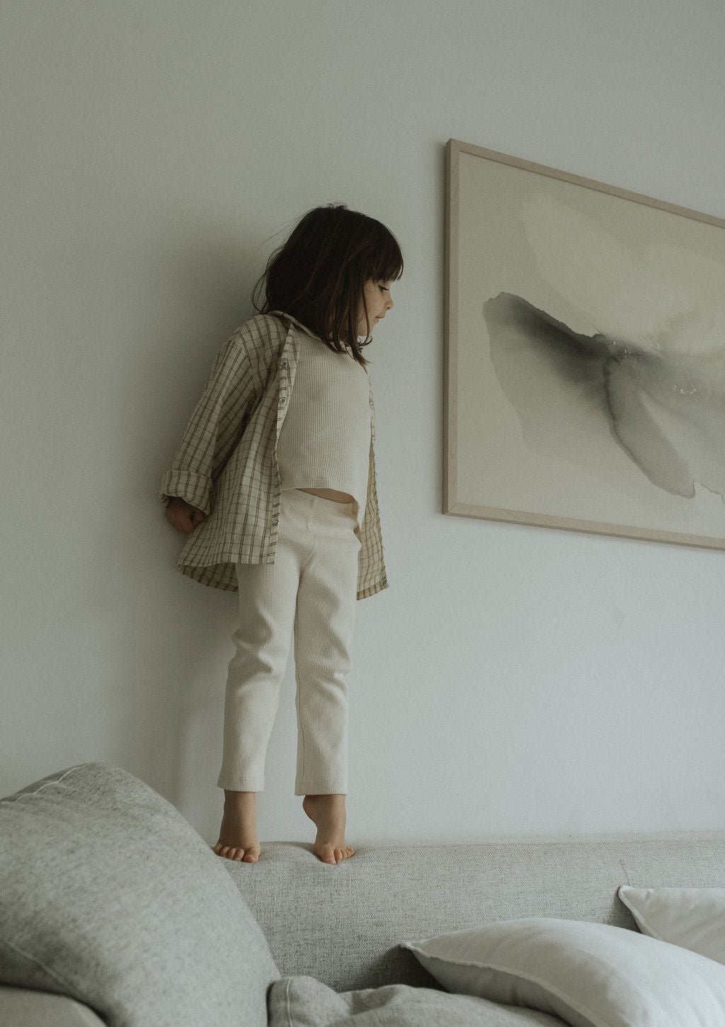 A young child in cabane childrenswear’s FAVORIS rib pant stands barefoot on a light gray couch. They wear a striped shirt, and a large neutral abstract painting hangs on the white wall behind them.