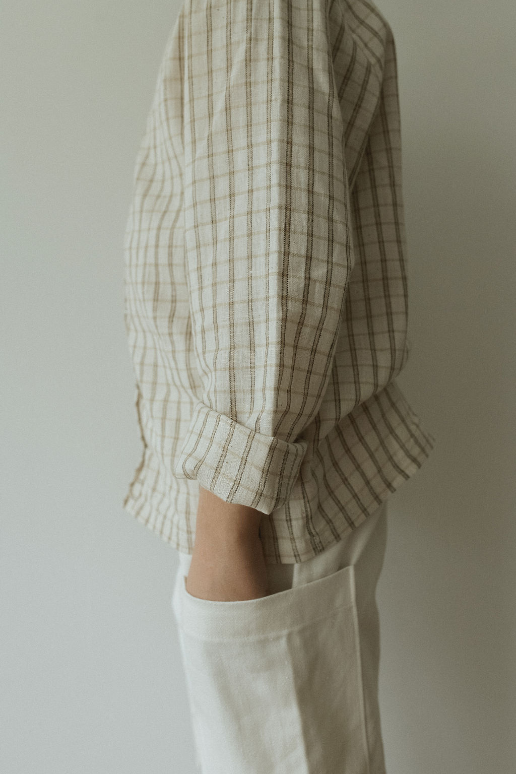 A person wears the cabane childrenswear TAKE CARE TOP in olive check, an ethically handmade long-sleeve shirt, with white pants, standing with one hand in their pocket against a neutral background.