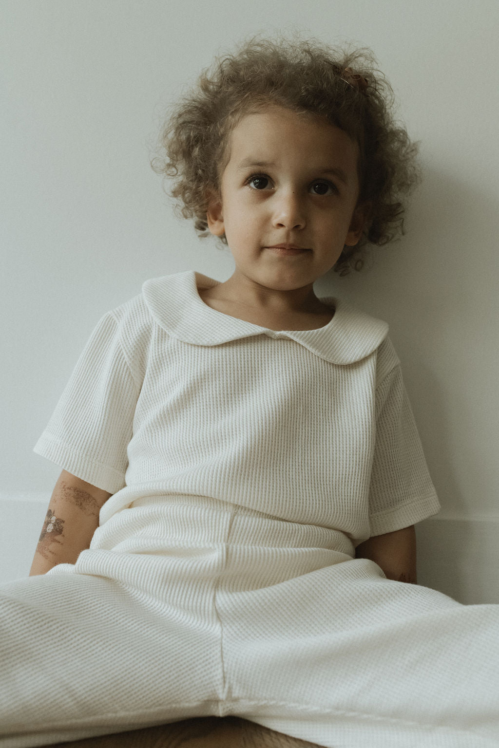 A curly-haired child sits against a white wall, wearing a SCHOOLHOUSE TEE waffle by cabane childrenswear. Calm and receptive, the child is seated on the wooden floor with legs apart.