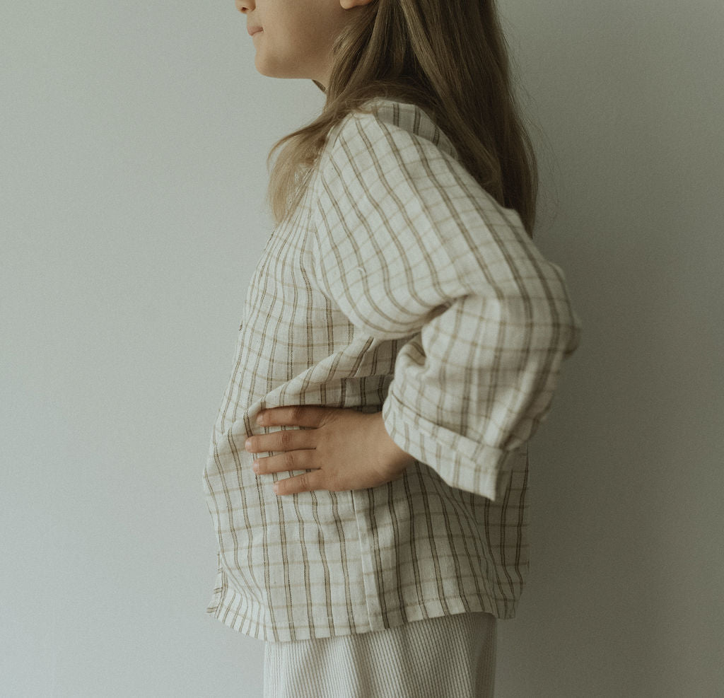 A person in profile against a light wall wears the TAKE CARE TOP in olive check by cabane childrenswear. The organic cotton plaid top features snaps, with one hand on their hip, face partly out of frame, and long hair loose.