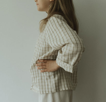 A person in profile against a light wall wears the TAKE CARE TOP in olive check by cabane childrenswear. The organic cotton plaid top features snaps, with one hand on their hip, face partly out of frame, and long hair loose.