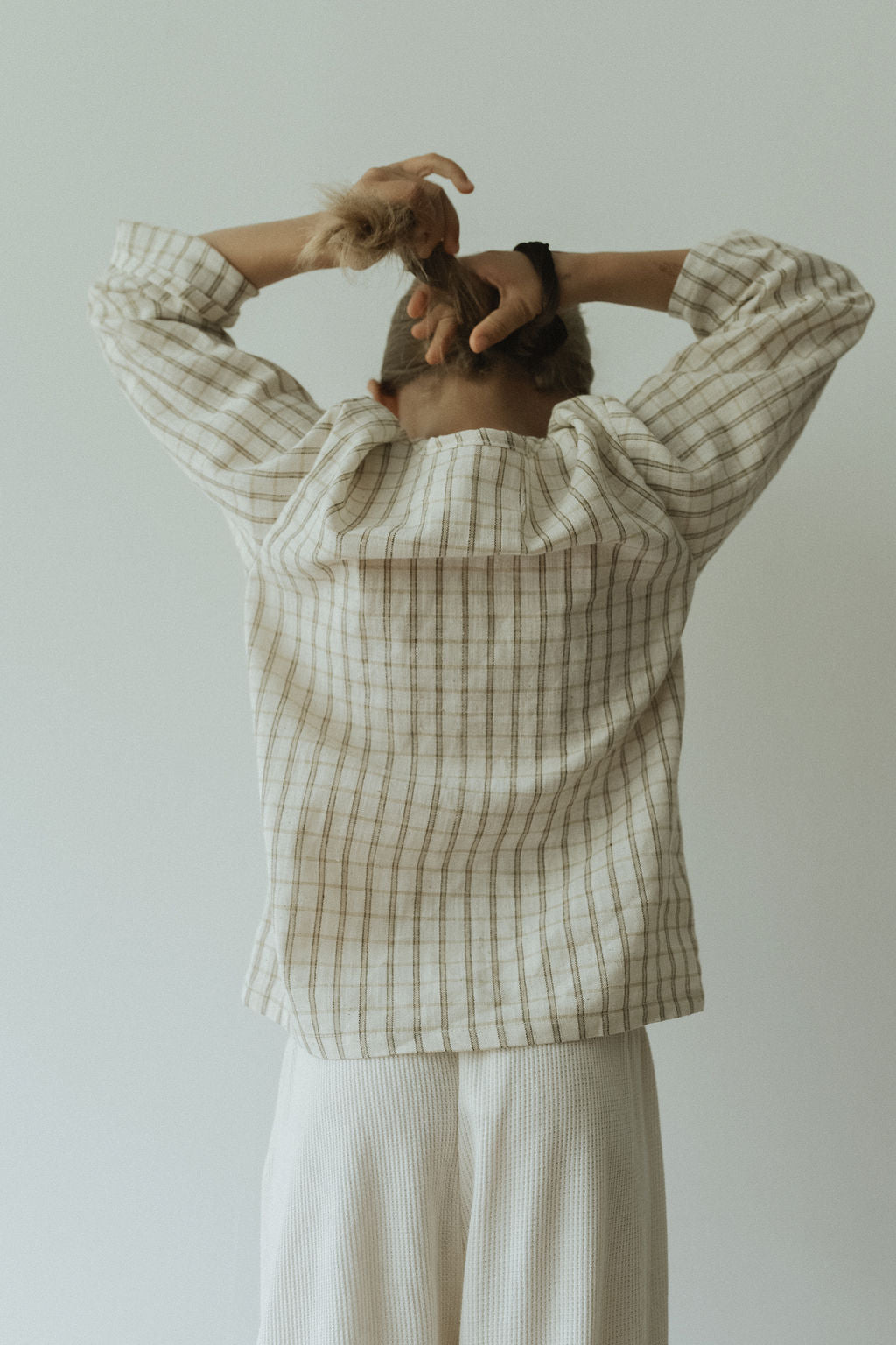 A person in the cabane childrenswear TAKE CARE TOP olive check, a loose, plaid, long-sleeve snap shirt made from soft organic cotton, pairs it with cream pants and ties their hair against a light background. Ethically handmade.