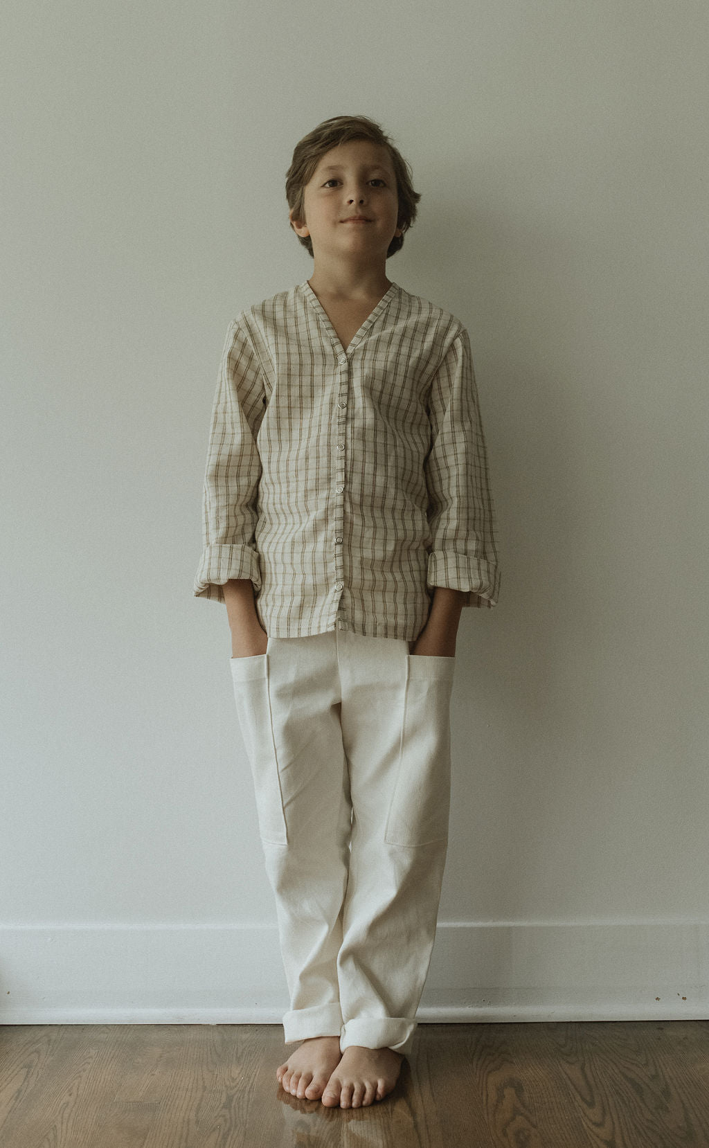 A young boy stands barefoot indoors against a plain wall, wearing the cabane childrenswear TAKE CARE TOP in olive check and white pants with his hands in his pockets.
