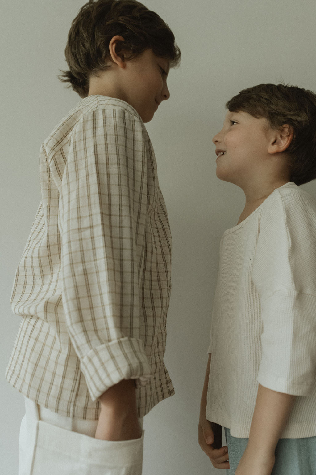 Two young boys face each other, smiling. One wears a beige plaid shirt and white pants; the other wears the cabane childrenswear TAKE CARE TOP in olive check. The neutral background highlights their joyful interaction.