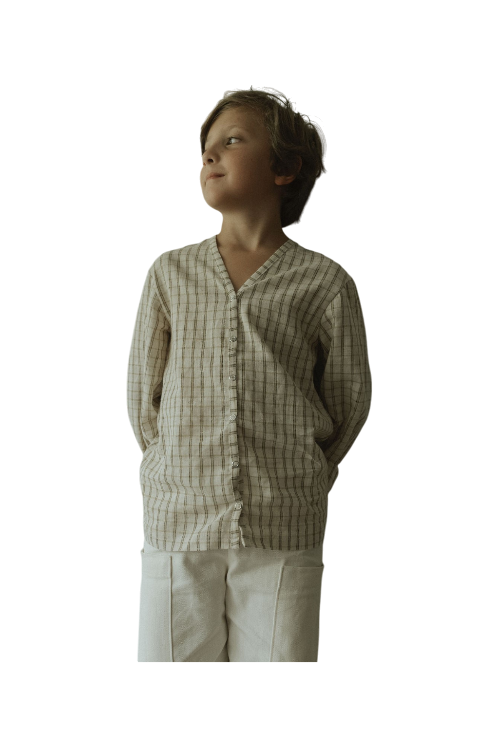 A young boy stands with his hands behind his back, smiling softly and looking to the side. He wears the cabane childrenswear TAKE CARE TOP in olive check. The background is transparent.