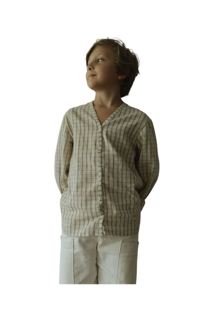 A young boy stands with his hands behind his back, smiling softly and looking to the side. He wears the cabane childrenswear TAKE CARE TOP in olive check. The background is transparent.