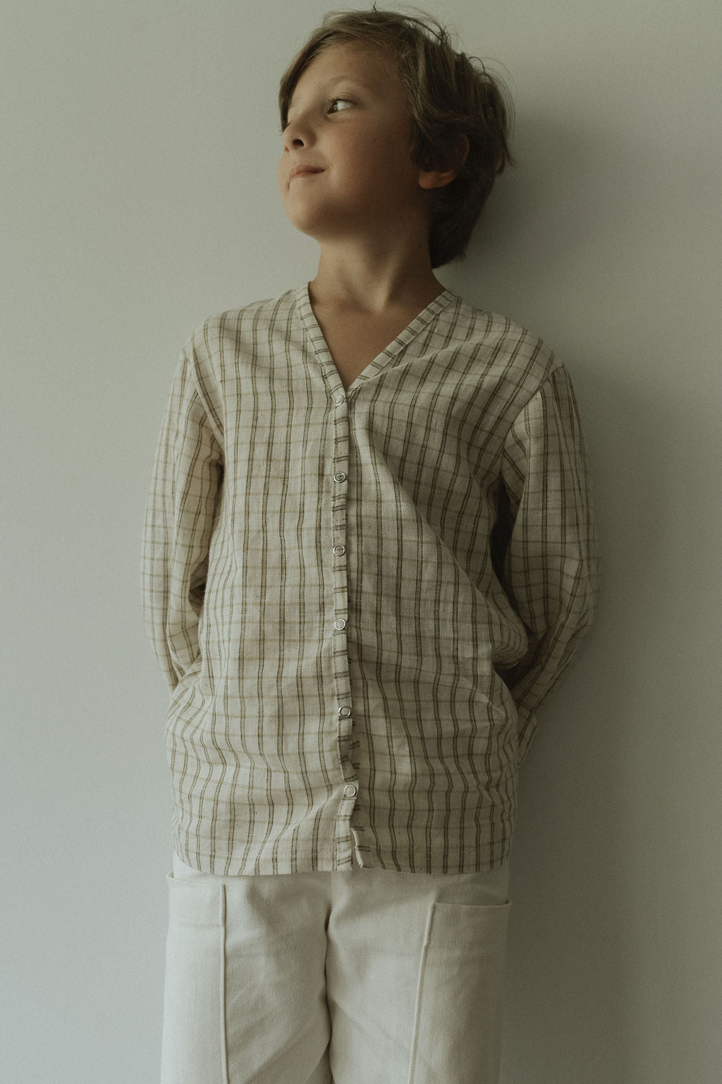 A young boy stands against a light wall, smiling softly. He wears the ethically handmade TAKE CARE TOP olive check by cabane childrenswear and light-colored pants, with his hands behind his back.