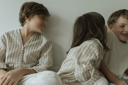 Three children sit together by a light wall. One whispers to another, who smiles, as the third looks on gently. They all wear cabane childrenswear’s TAKE CARE TOP olive check, crafted from soft organic cotton in light, neutral tones.