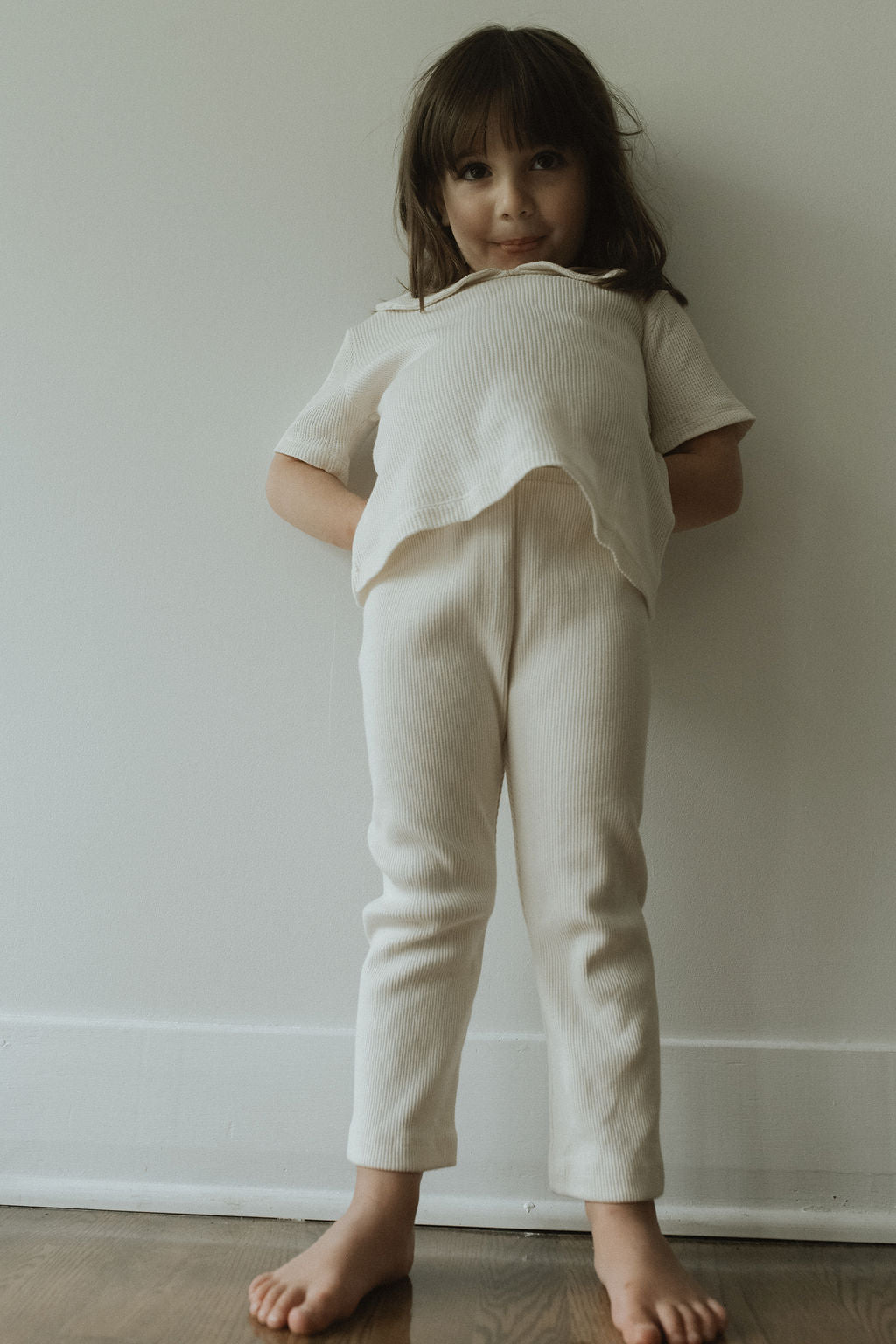 A young girl with long brown hair stands barefoot indoors against a white wall, wearing cabane childrenswear: a light-colored FAVORIS rib pant paired with a matching ribbed cotton top, smiling softly with her hands tucked behind her back.