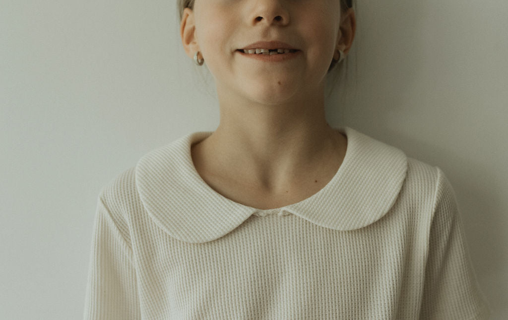 A young girl stands against a light background, embodying classic looks in Cabane childrenswear's SCHOOLHOUSE TEE waffle with a Peter Pan collar. Her hair is tied back, and her slight smile reveals grace and timeless charm.