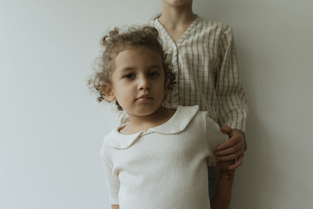 A young child with curly hair wears the SCHOOLHOUSE TEE waffle from cabane childrenswear, standing in front and looking at the camera. Behind them, another person in a plaid shirt gently rests a hand on their shoulder against a plain white backdrop, enhancing their classic look.
