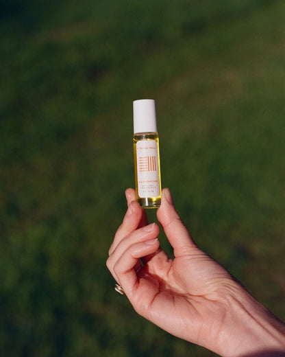 A hand displays & Tread Well’s Natural Fragrance – Illumination, a roll-on perfume with yellow liquid, white cap, and minimalist label—providing natural aromatherapy for stress relief amid a blurred green outdoor backdrop.