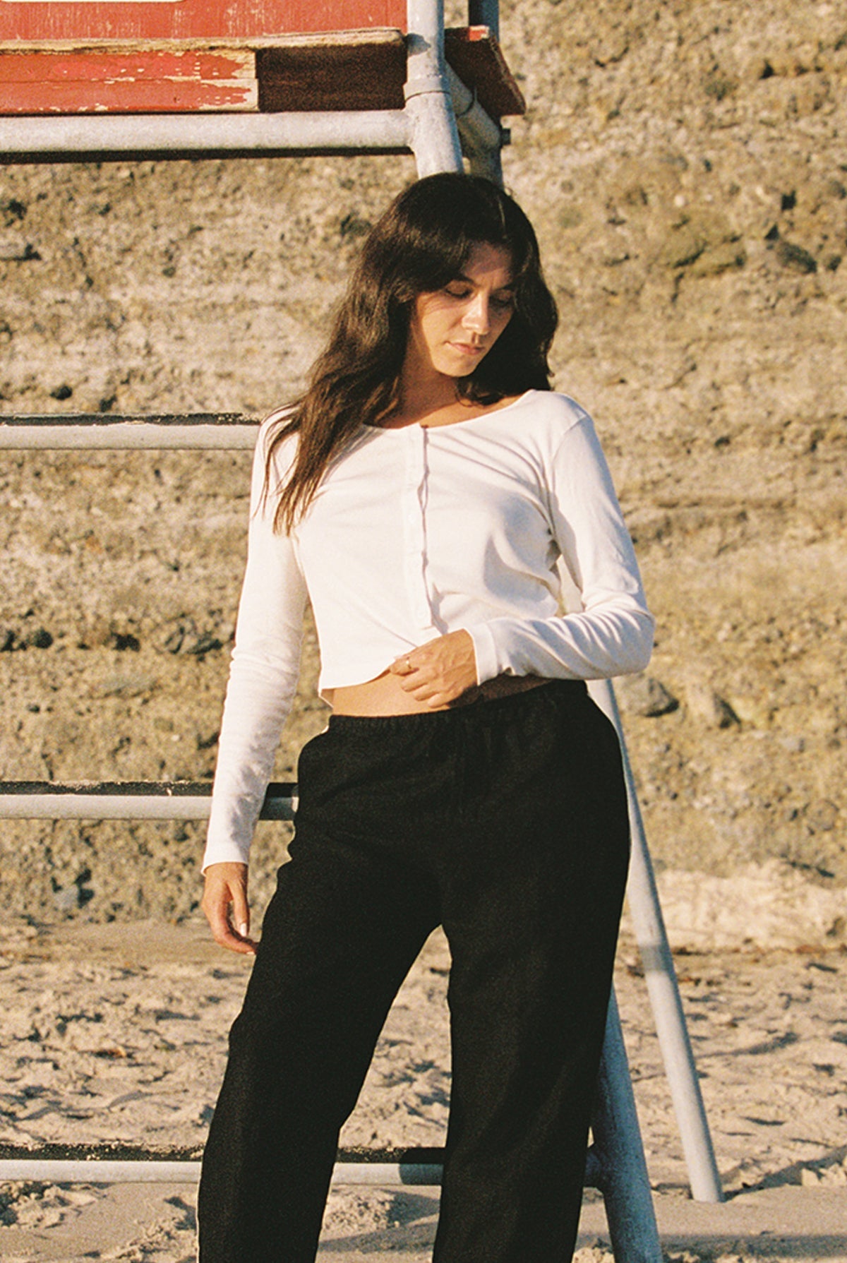 Ivory - A woman in an Organic Rib Cardigan by LA Relaxed and dark pants stands beside a metal ladder on the sunlit beach. Her long brown hair cascades as she looks down, with a rocky wall behind her, channeling effortless 90's style.